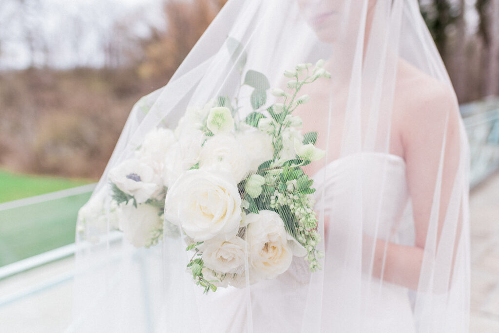 A bride in a strapless white gown holds a bouquet of white garden roses, ranunculus, anemones, and greenery, softly veiled by sheer tulle in the foreground, with a blurred autumn landscape in the background.