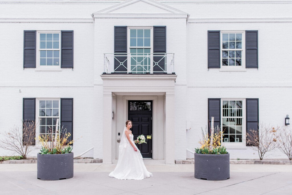 Bride in a white gown standing before a symmetrical white Georgian building with navy shutters and a balcony, Harding Waterfront Estate (Burlington), a timeless Old World European wedding venue in Canada.