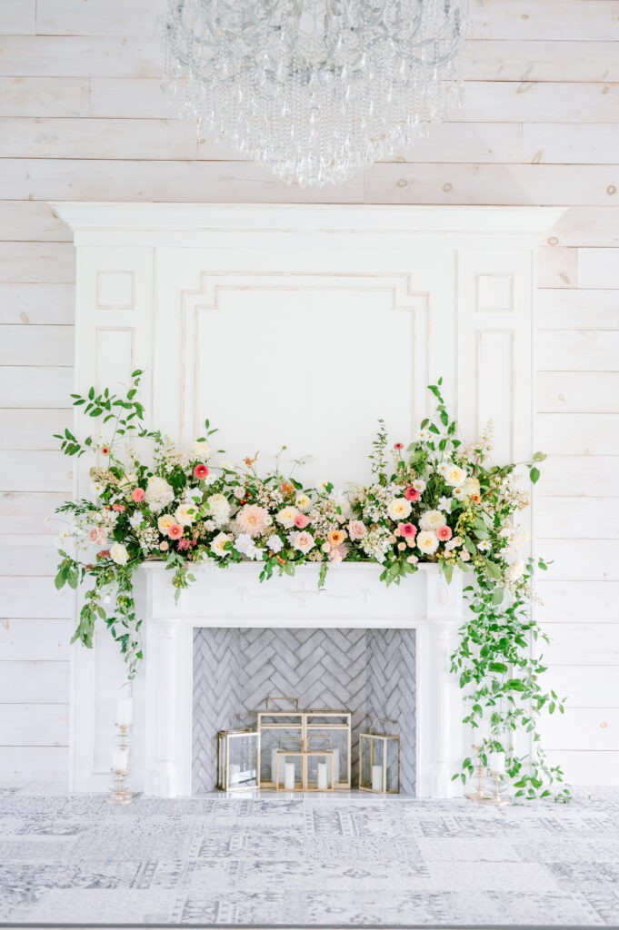 White fireplace mantel at Stonefields Estate decorated with a lush garland of pink and cream florals, with candle lanterns inside the herringbone-tiled hearth.