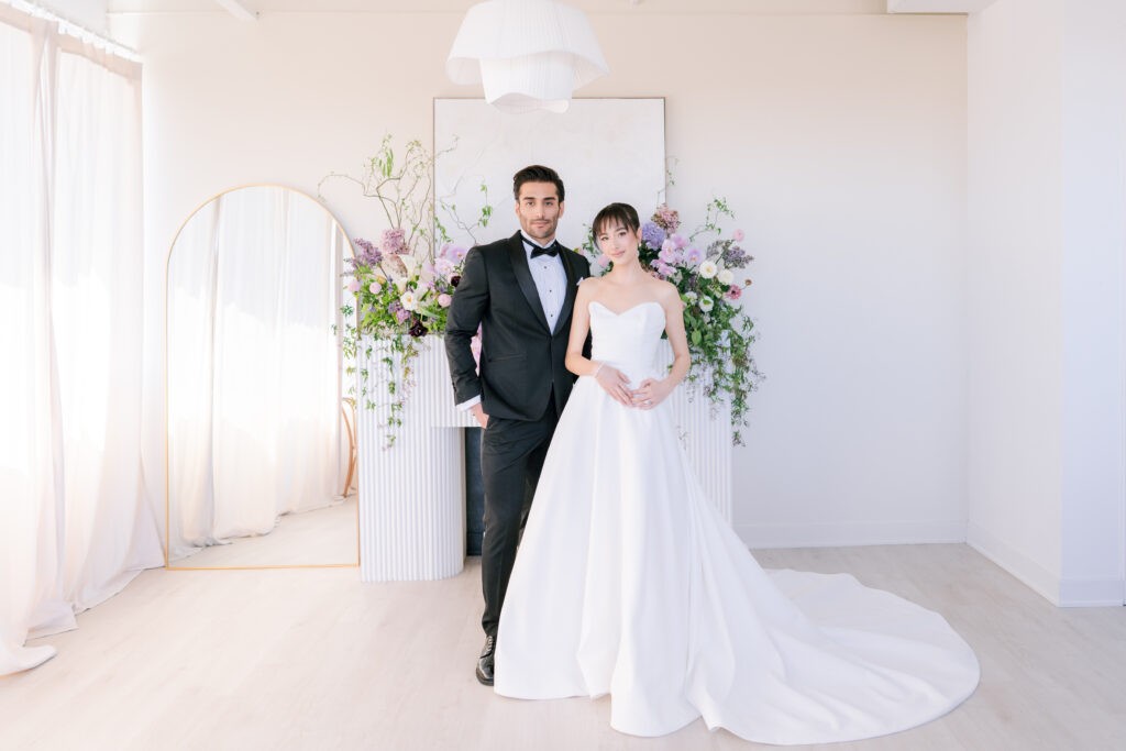 Bride from White Satin Bridal wearing a sweetheart-neckline ball gown with a long train, standing with a groom in a black tuxedo in front of a floral display and arched mirror in a bright, minimalist room
