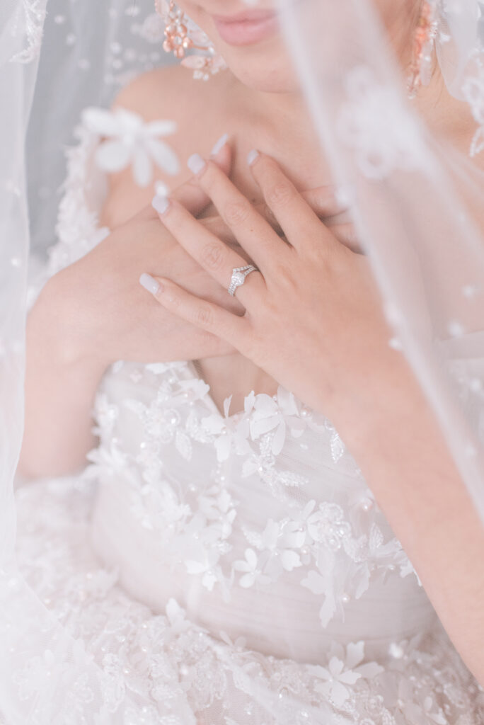 Close-up of a bride from Fairy Dreams Bridal touching her shoulder, showcasing a floral appliqué gown, diamond ring, and delicate lace veil against a pink backdrop