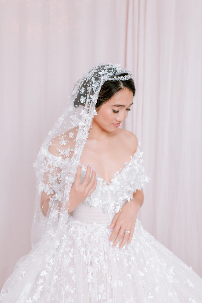 Bride from Fairy Dreams Bridal in an off-shoulder floral lace ball gown, veil draped over her head and face, standing against a soft pink curtain backdrop