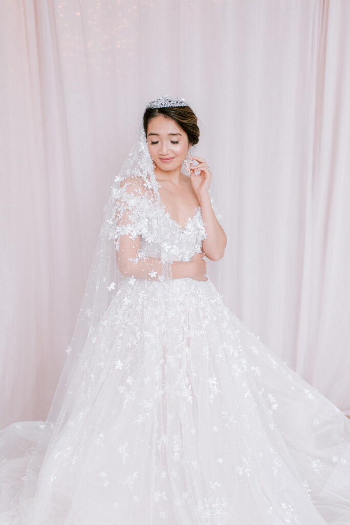 Bride from Fairy Dreams Bridal wearing a floral lace ball gown with a plunging neckline, tiara, and sheer veil, looking down softly against a pink curtain backdrop