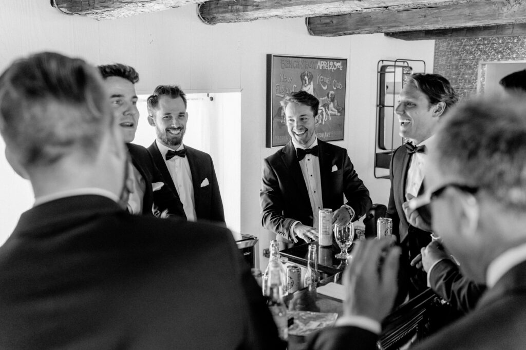Black and white photo of groomsmen in tuxedos laughing together around a drinks table at Stonefields Estate, with a vintage poster on the wall behind them.