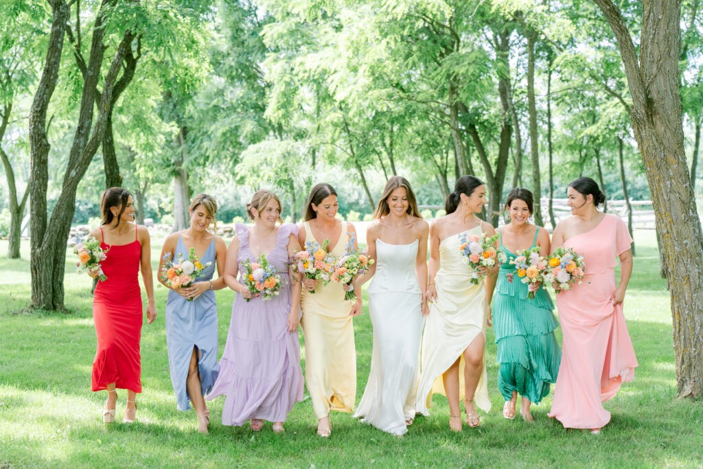 Bride walking with bridesmaids in mismatched pastel and jewel toned dresses, each carrying a colorful floral bouquet, on a sunlit lawn.