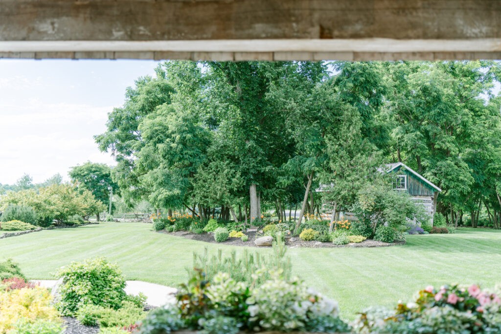 View of manicured gardens and lawns at Stonefields Estate, framed by a wooden beam, with a small outbuilding tucked among the trees.