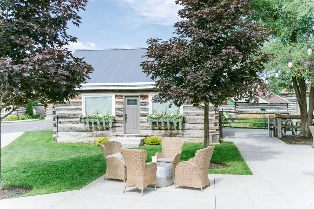 Wicker seating area outside a log cabin building at Stonefields Estate, shaded by maple trees on a paved patio.