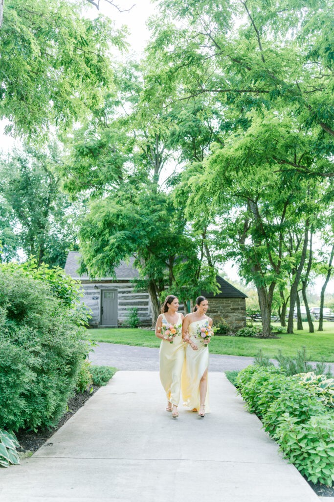 Bridesmaids in yellow dresses walking down a garden path lined with greenery at Stonefields Estate.
