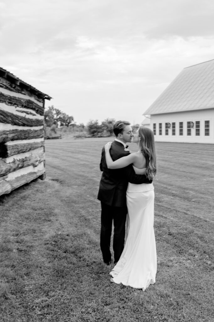 Black and white photo of a bride and groom embracing on the open lawn at Stonefields Estate, with a log cabin and chapel in the background.