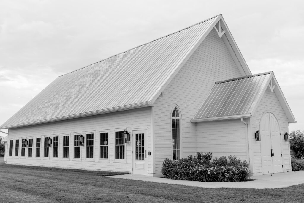 Black and white exterior photo of a white chapel-style building at Stonefields Estate, featuring a steep metal roof, arched window, and double entry doors framed by greenery.