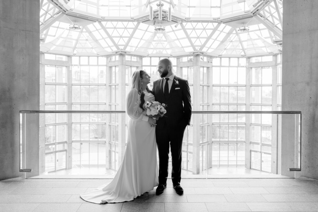 Black-and-white portrait of a bride and groom gazing at each other on a mezzanine inside the National Gallery of Canada, framed by the building's signature geometric glass rotunda.