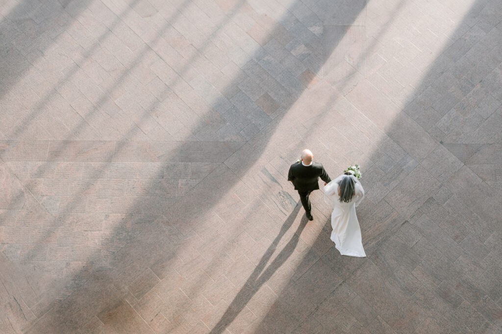 Aerial view of a bride in a white gown and veil and her groom in a black suit walking hand in hand across a sunlit stone plaza at the National Gallery of Canada in Ottawa.