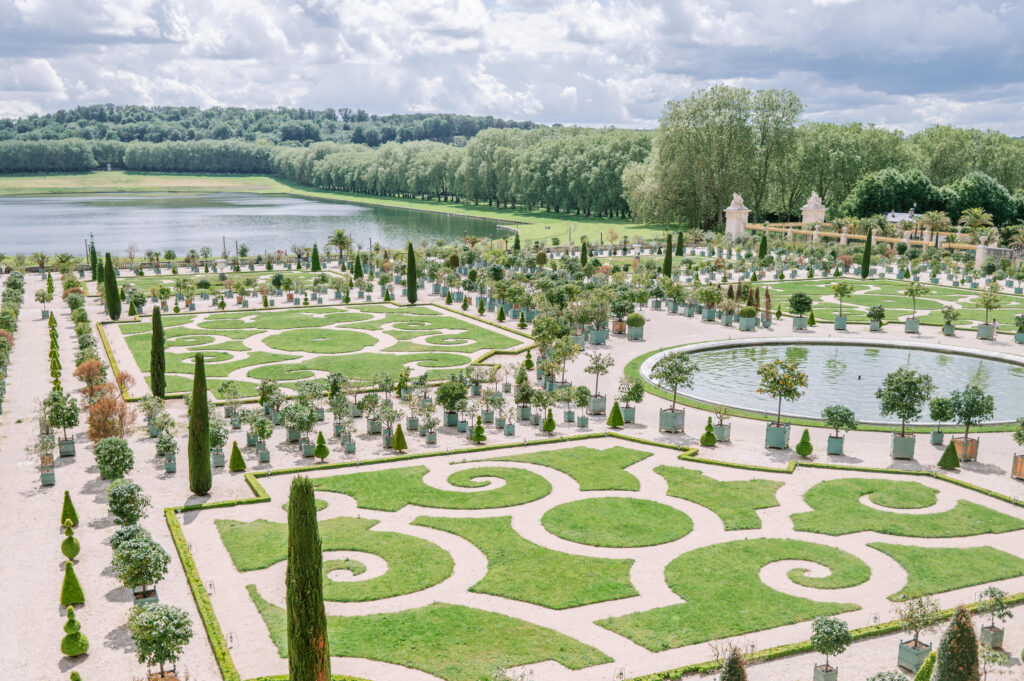 Elegant formal garden with intricate symmetrical patterns and spirals of green grass, surrounded by potted trees, set against a serene lake and lush forest backdrop.