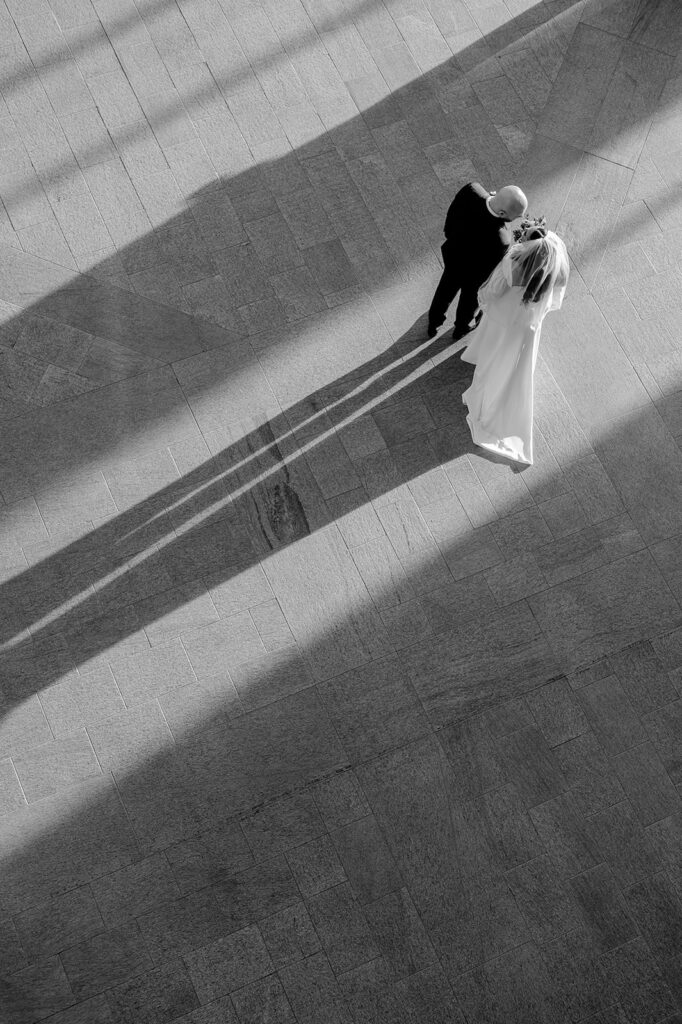 Aerial black-and-white photo of a bride and groom embracing on a tiled floor, their long shadows stretching across sunlit pavement.