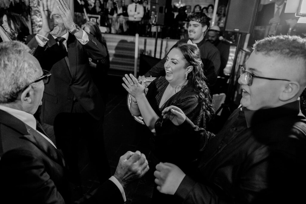 Black and white photo of guests dancing and clapping joyfully on the dance floor at a wedding reception.