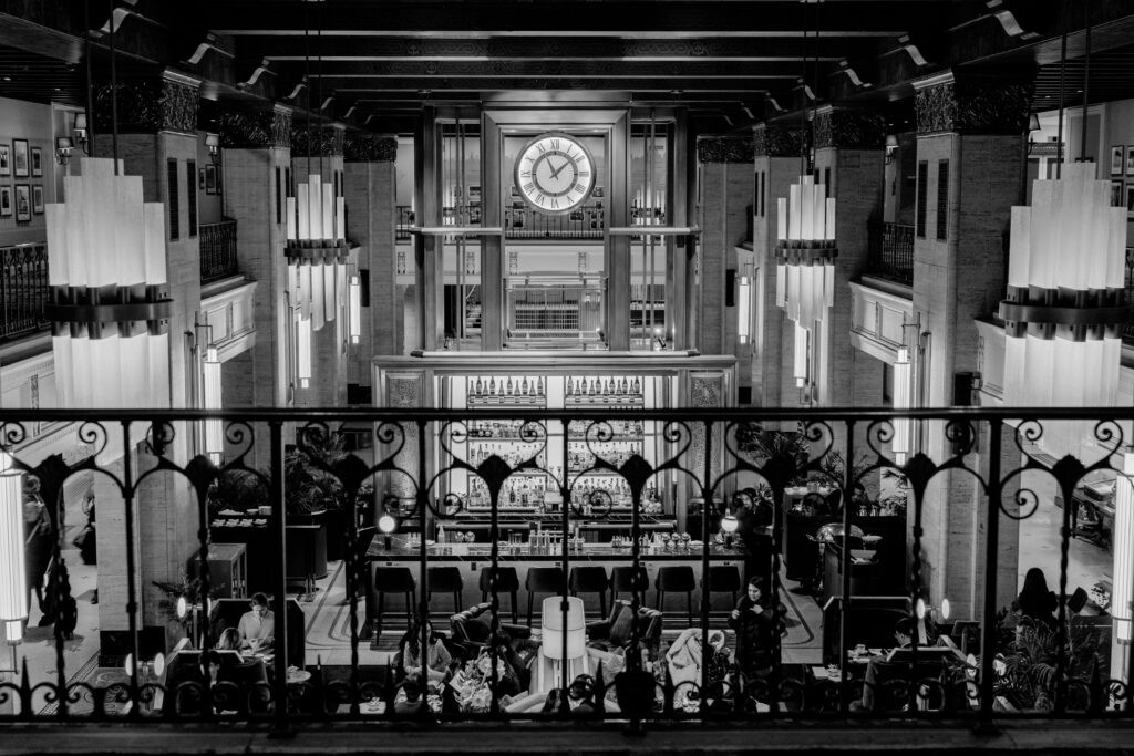Grand Art Deco hall with ornate wrought-iron balcony railing, tall pendant lights, and a large central clock over the bar — an Old World European wedding venue in Canada.