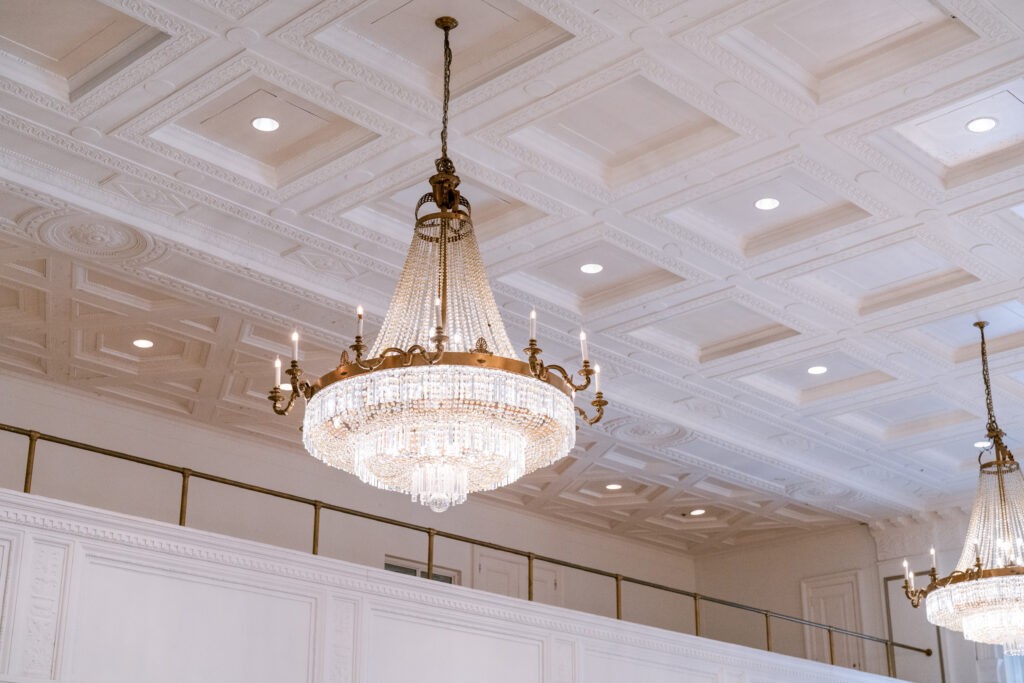 Crystal chandelier with brass candle arms hanging from a coffered ceiling at the Fairmont Royal York, with a matching chandelier and gold balcony railing visible in the background.