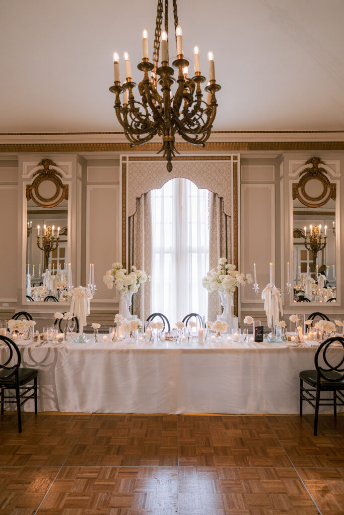 Head table reception setup framed by ornate mirrored paneling and an arched window, with a wrought-iron chandelier and floral arrangements above white place settings.