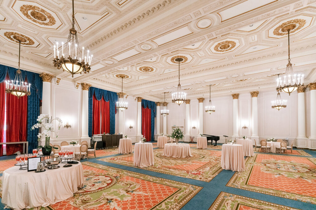 Grand cocktail hour setup in the Fairmont Château Laurier's Laurier Room, with ornate gilded ceiling coffers, brass chandeliers, rich blue and red draperies, and blush-linened cocktail tables on a patterned carpet.