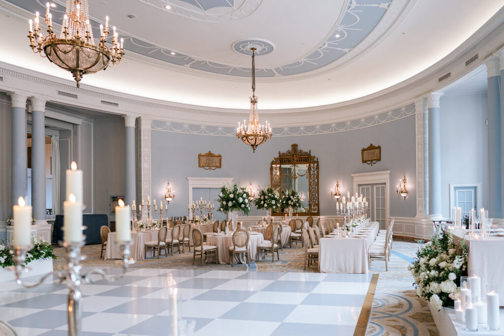 Elegant curved reception setup in the Fairmont Château Laurier's Adam Room, featuring brass chandeliers, a checkered marble dance floor, ornate mirrored fireplace, and candlelit tables with white floral arrangements, styled by Erica Irwin.