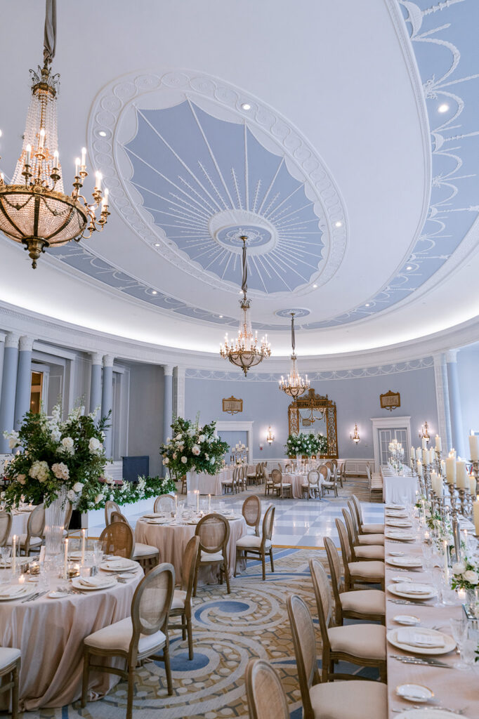 Wide view of the Adam Room's curved reception layout, with rattan-back chairs, taper candles, and floral centerpieces set beneath ornate plasterwork and chandeliers, styled by Erica Irwin.