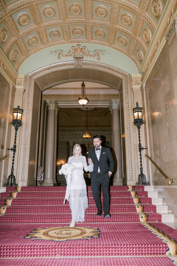 Bride and groom descending a red-carpeted staircase beneath an ornate painted archway at the Fairmont Château Laurier.