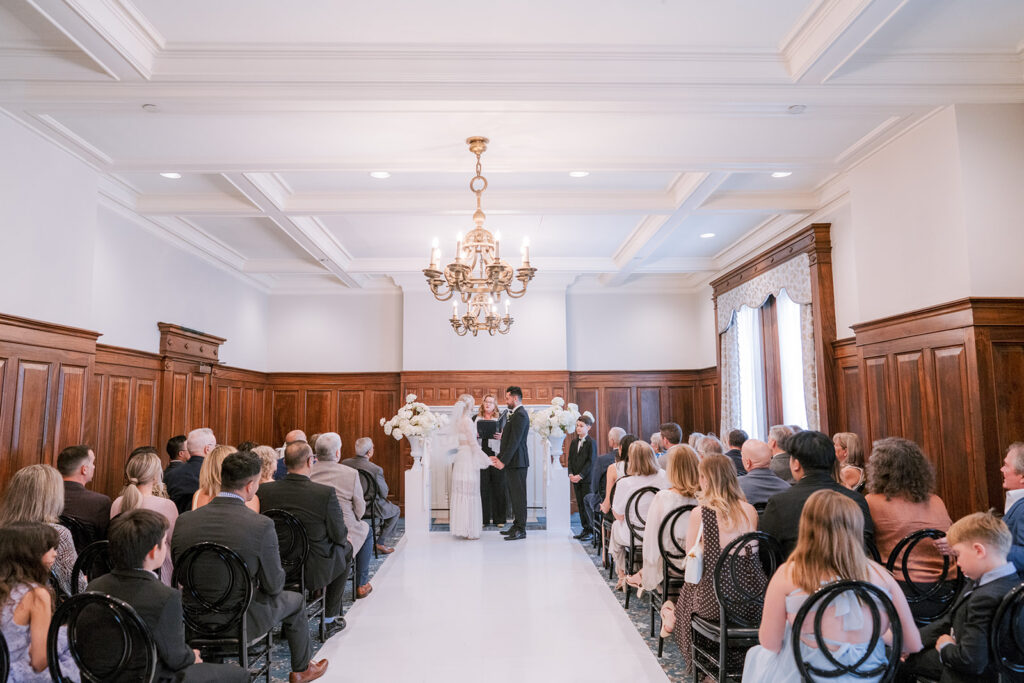 Wedding ceremony at the Fairmont Château Laurier, with the couple exchanging vows beneath a brass chandelier, flanked by wood-paneled walls and seated guests.