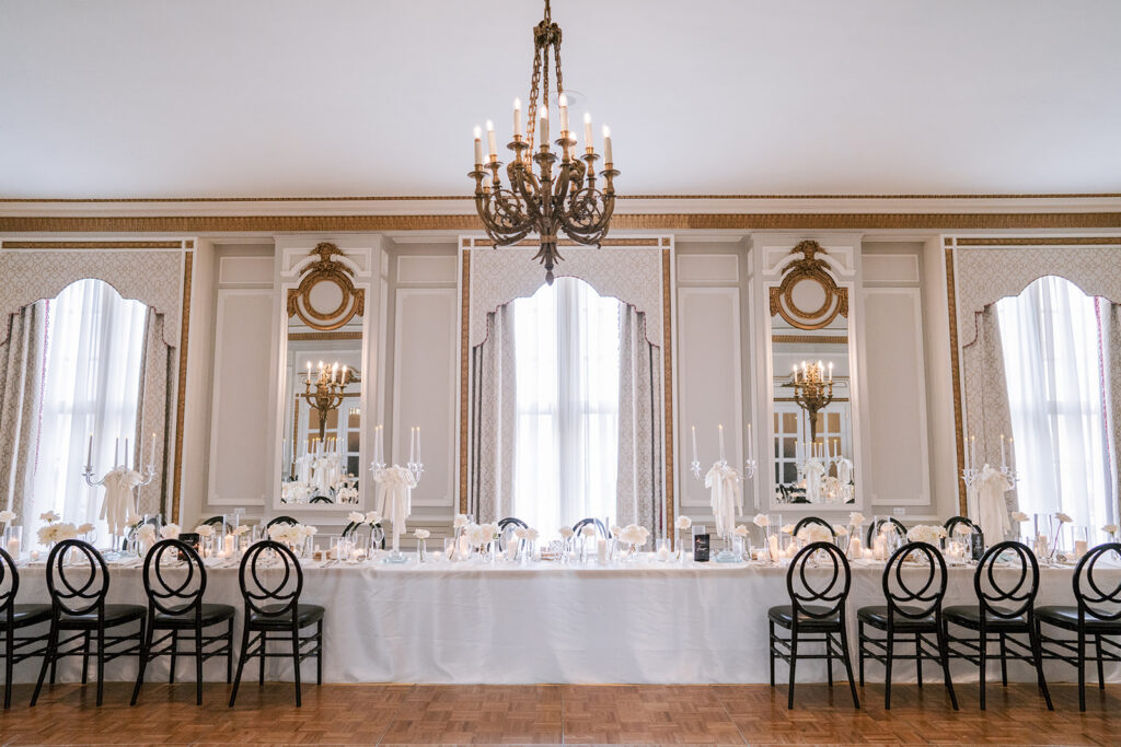 Head table wedding reception setup at the Fairmont Château Laurier, framed by mirrored wall panels and arched draped windows, with black wishbone chairs and a brass chandelier overhead.