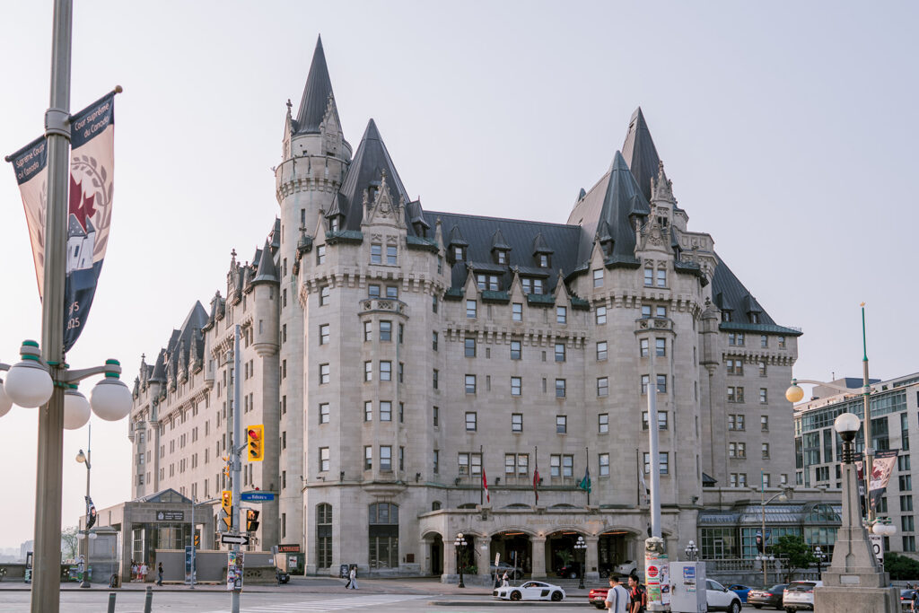 Exterior street view of Fairmont Château Laurier in Ottawa, a grand château-style hotel with turreted towers and steep copper roofs, framed by city lamp posts and passing traffic.