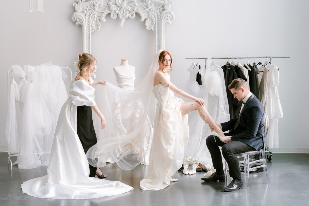 Bride getting ready in a bridal boutique, with a groom kneeling to help with her shoe and a stylist holding her train, surrounded by dress racks and an ornate white mirror.