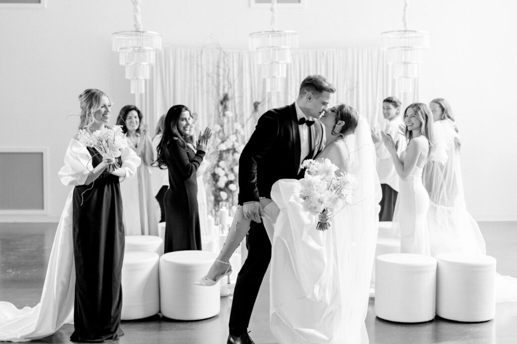 Black and white photo of a groom dipping his bride for a kiss during their first dance, surrounded by cheering guests and bridesmaids, with tiered crystal chandeliers and a floral backdrop.