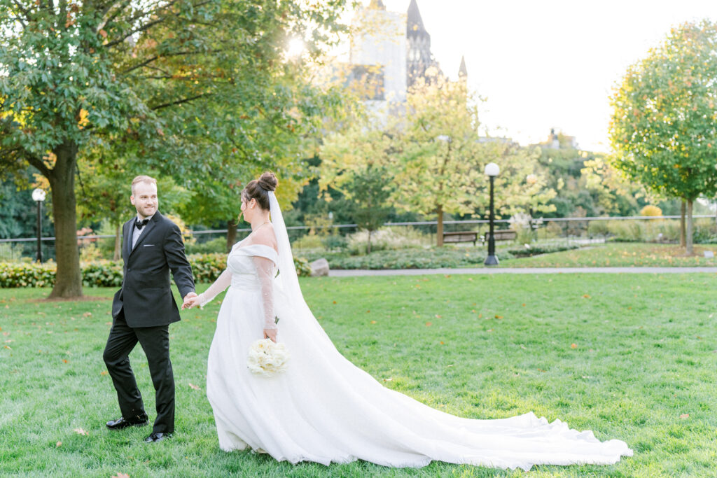 Groom leading the bride by the hand across a sunlit park lawn, her long train trailing behind, with château style towers in the background.