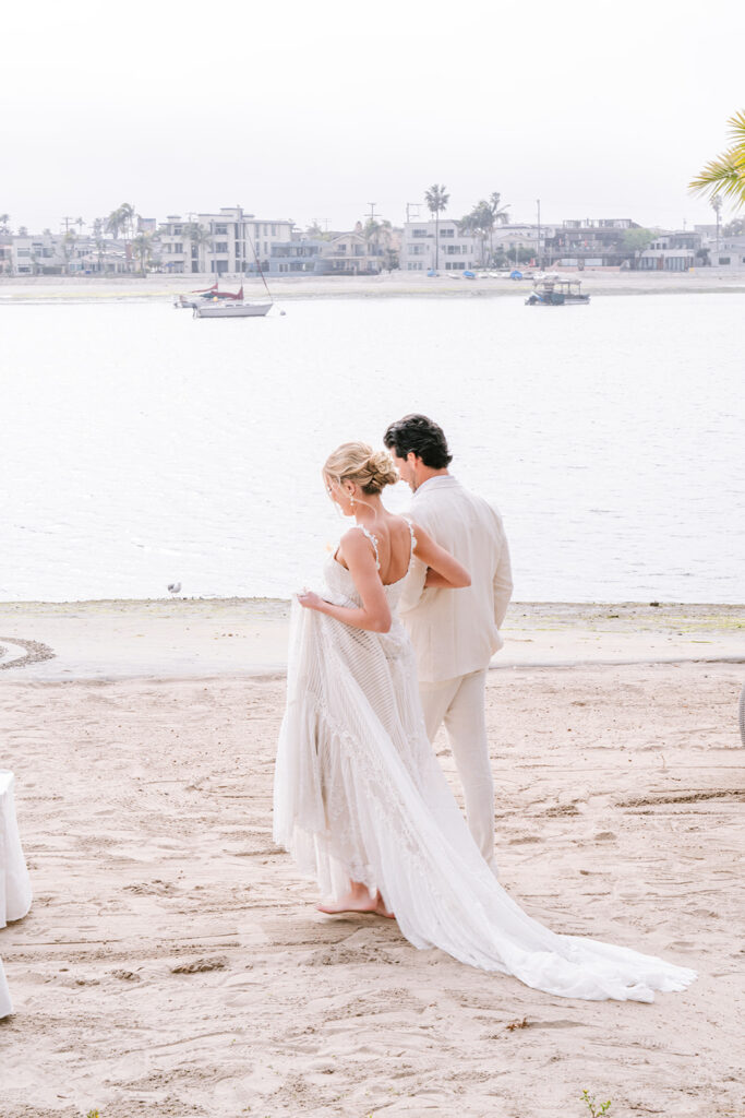 Bride in a flowing lace gown walking barefoot on the sand with the groom beside a calm bay, seaside wedding at San Diego, California.