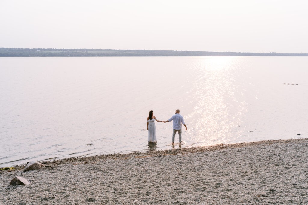 Couple holding hands while wading into a calm lake at sunset, with a sandy shoreline in the foreground.