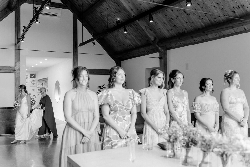 Black and white photo of bridesmaids in patterned dresses standing in a row inside a wood beamed barn venue, as the bride walks in behind them.