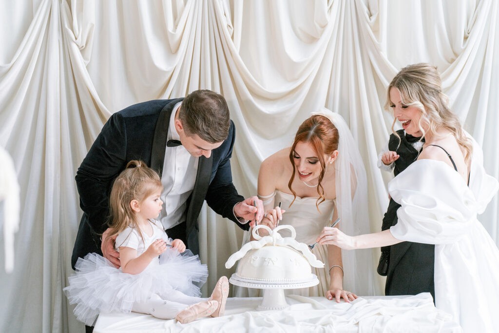 Bride and groom cutting a white bow topped wedding cake together with a young flower girl in a tutu and a guest looking on, in front of draped fabric backdrop.