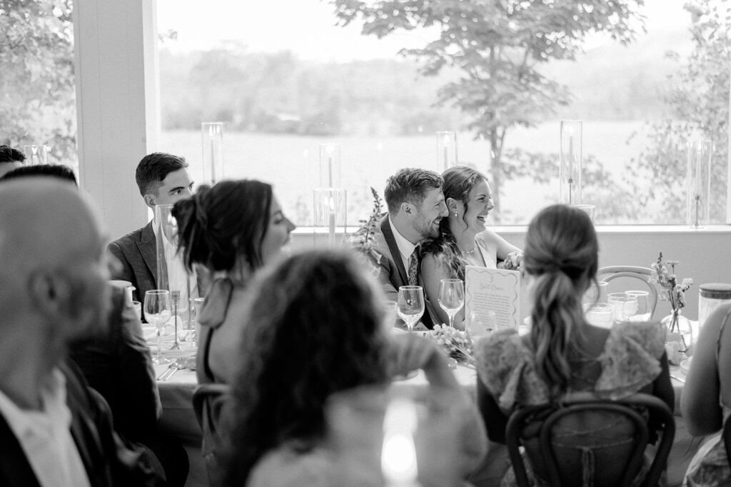 Black-and-white photo of a wedding reception. A bride and groom sit at the head table, laughing together, surrounded by seated guests in the foreground.