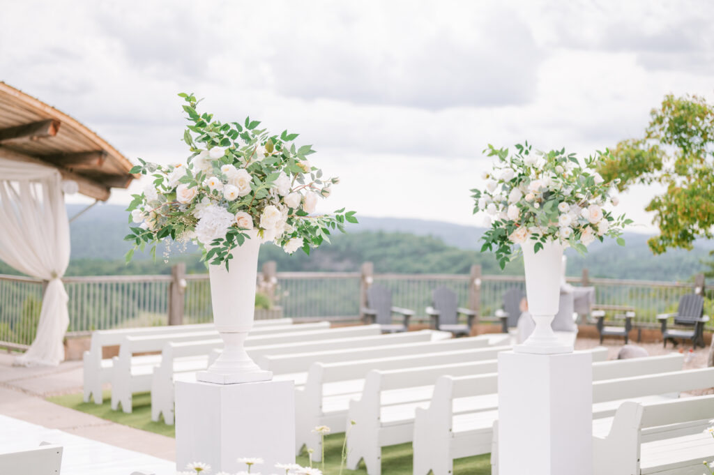 White floral arrangements on tall pedestals lining the outdoor ceremony aisle at Le Belvédère, with white benches and a scenic mountain backdrop.