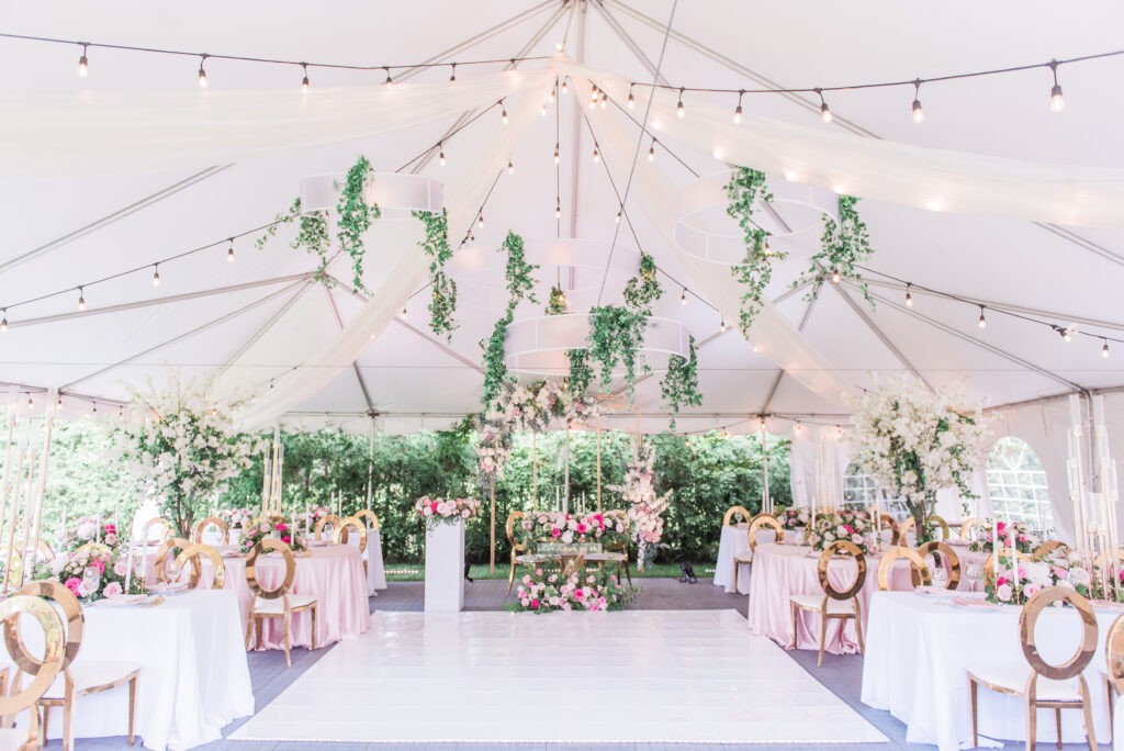 Wide view of a tented celebration at a private estate, featuring a white dance floor, hanging greenery installations, string lights, and pink and gold table settings.