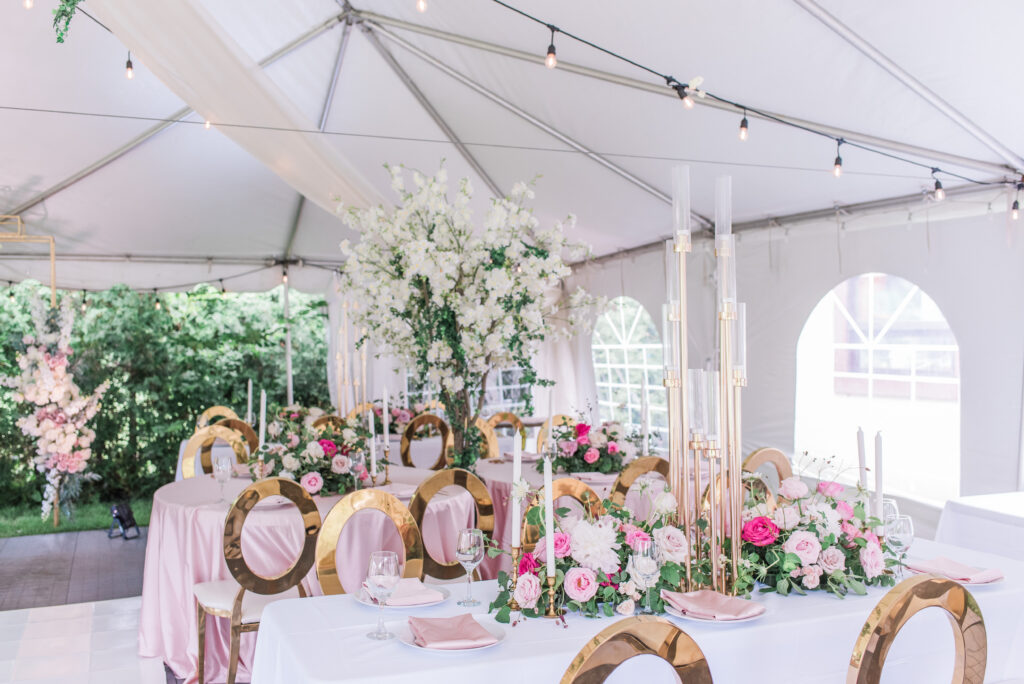 Tented reception at a private estate wedding, with tall white floral arrangements, gold candlesticks, blush linens, and circle-back chairs under string lights.