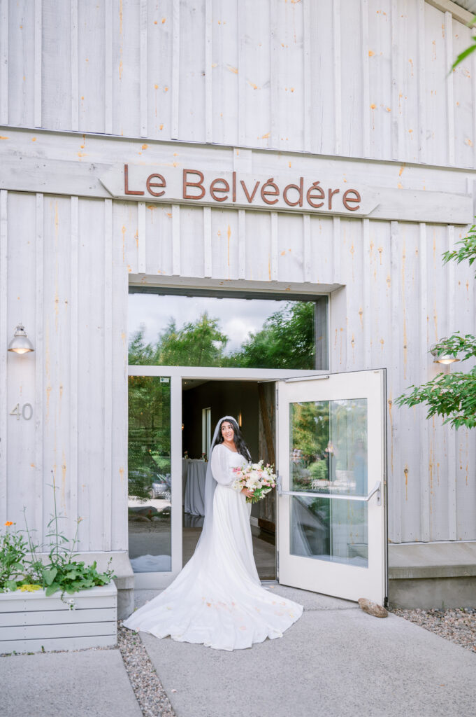 Bride standing in the doorway of Le Belvédère, a rustic white wood venue with wooden signage and a bouquet in hand.