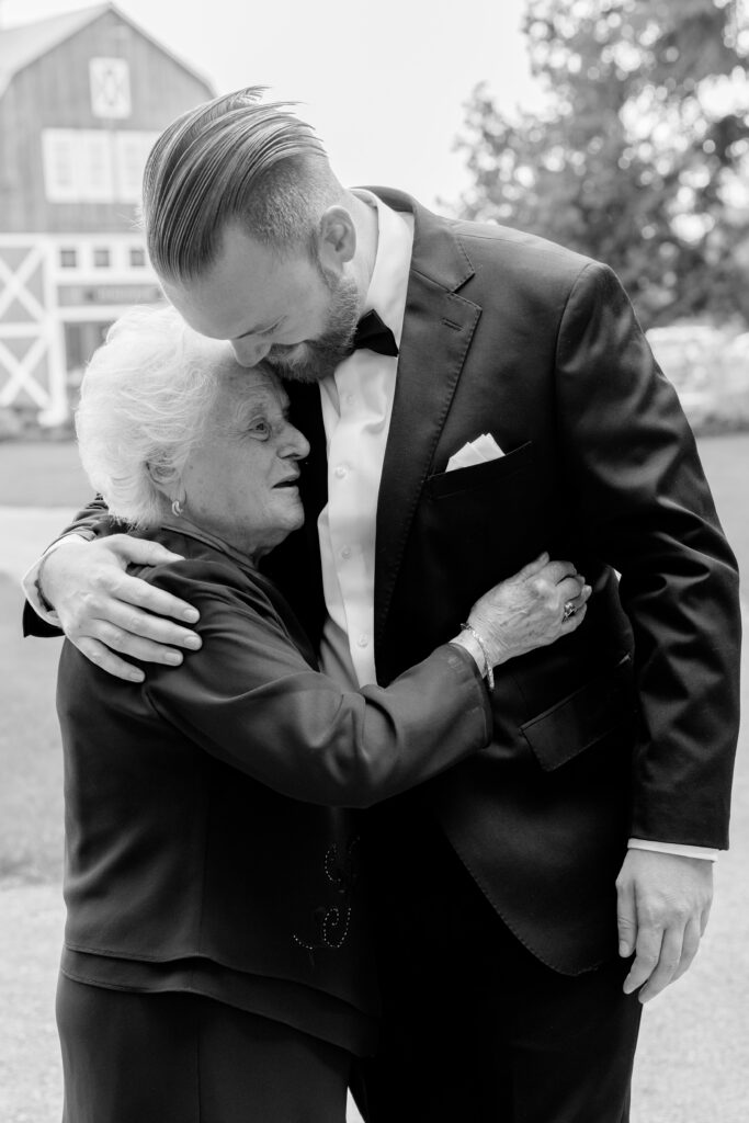 Black-and-white photo of a groom embracing an older woman warmly outdoors, with a red barn visible in the background.