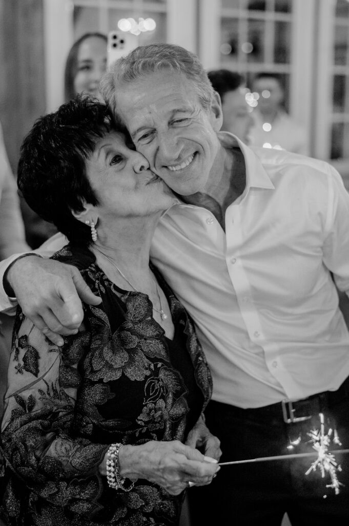 Black-and-white photo of a man affectionately kissing an older woman on the cheek while she holds a sparkler at a wedding celebration.