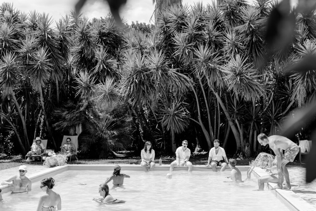 Black and white photo of guests relaxing and swimming in a pool surrounded by tall palm trees, part of a wedding weekend in Italy.