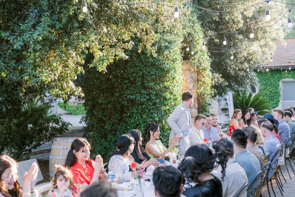 Guests seated at a long outdoor reception table clapping as a man gives a toast, string lights and greenery overhead, during a wedding weekend in Italy.