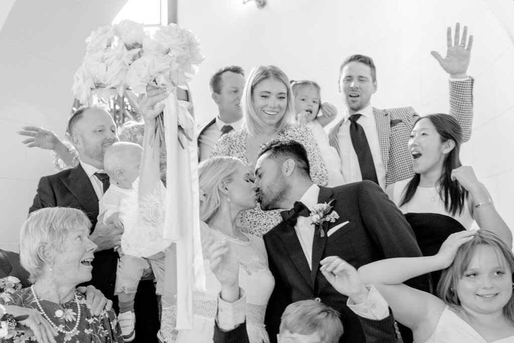 Bride and groom kiss while family and friends celebrate around them during a joyful wedding portrait.