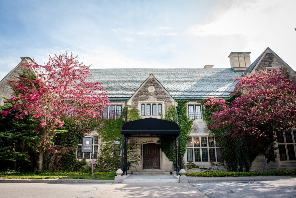 Ivy covered stone manor with steep gabled roofs and blossoming pink trees framing the entrance, Estates of Sunnybrook (Toronto), a stately Old World European wedding venue in Canada.