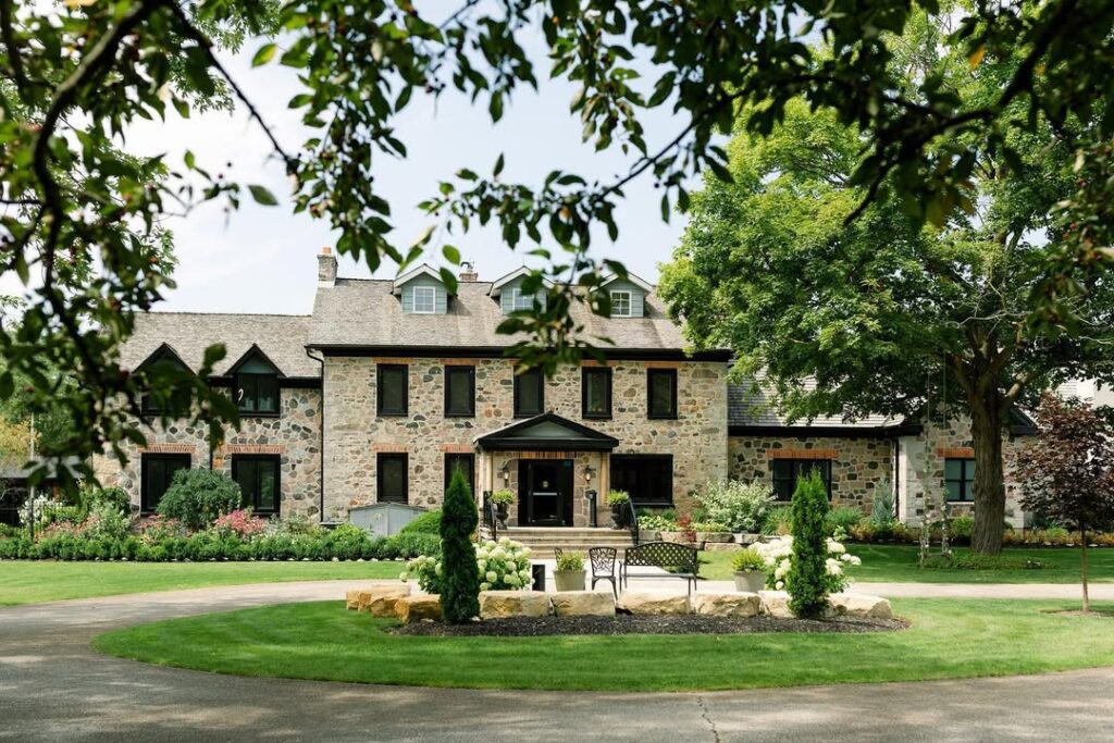 Stone farmhouse style estate framed by tree branches, with a circular driveway, manicured lawn, and garden beds, Roseville Estates (Cambridge), a charming Old World European wedding venue in Canada.