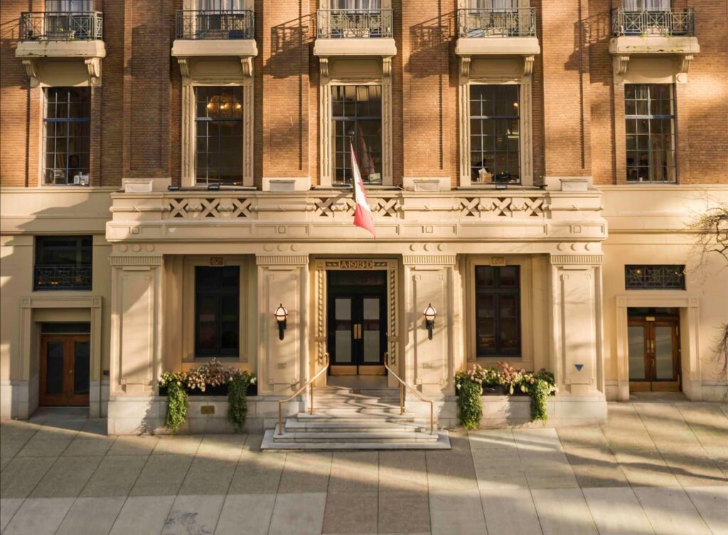 Golden-hour exterior of The Vancouver Club, a brick and stone heritage building with wrought-iron balconies, flower boxes, and a Canadian flag hanging above the entrance.