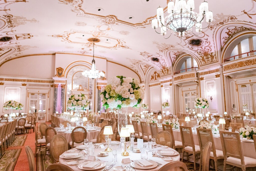 Opulent ballroom wedding reception in Quebec City with ornate gilded ceiling frescoes, crystal chandeliers, tall white floral centerpieces, and rattan-back chairs at round tables.